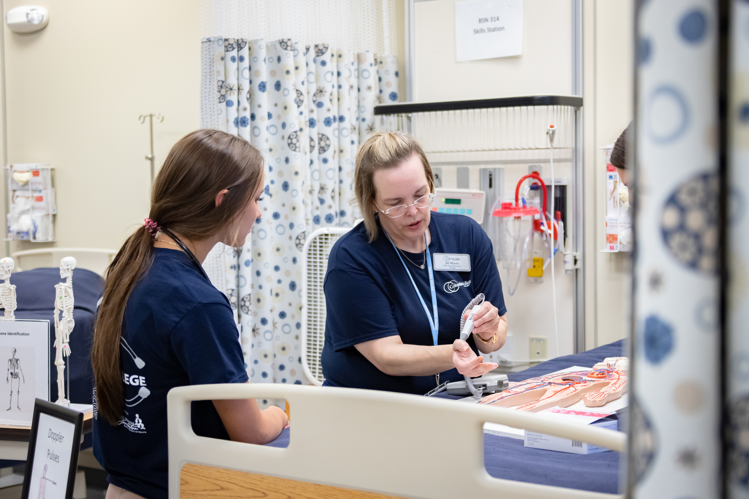 Two nursing students standing around a "patient" bed, one running tests while the other watches.