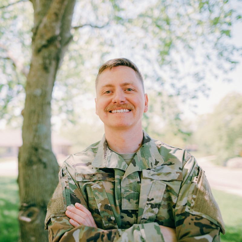 Military: Man with arms crossed wearing military attire while standing in front of a tree.
