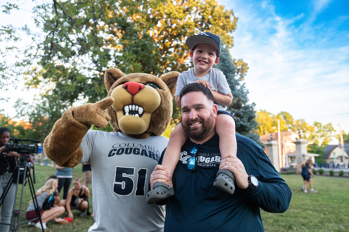 A father, with his young son on his shoulders, stands next to Scooter the Cougar on the Columbia College main campus.