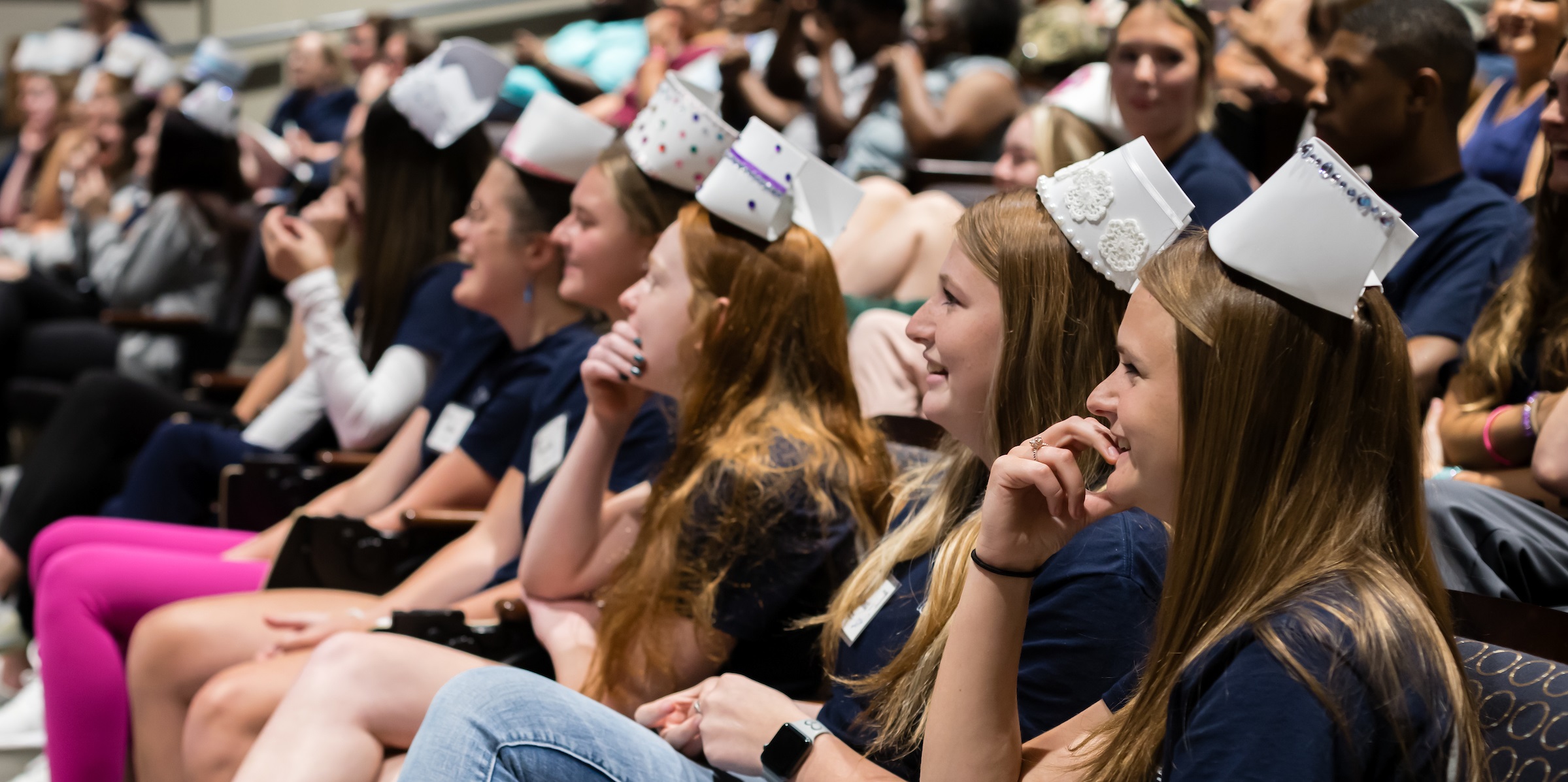 Group of nursing students sit in auditorium wearing their homemade nursing caps.
