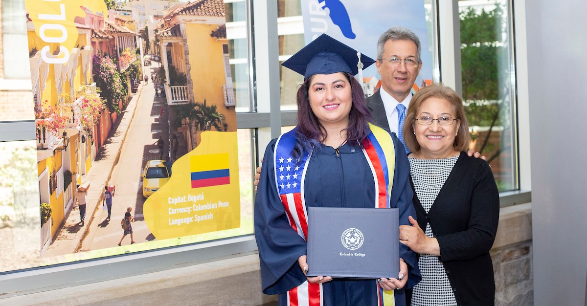 Graduate, in full regalia, standing her diploma and standing next to her mom and dad in front of a poster about Bogota.