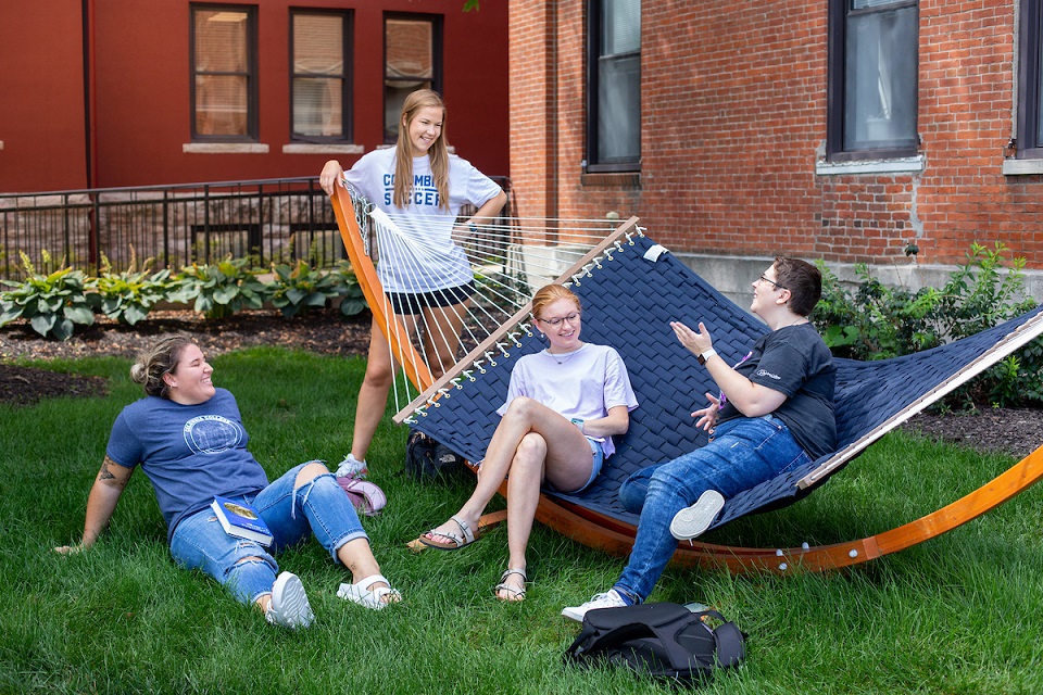 Four students outside engaged in conversation, with two sitting on one of the many hammocks on main campus.