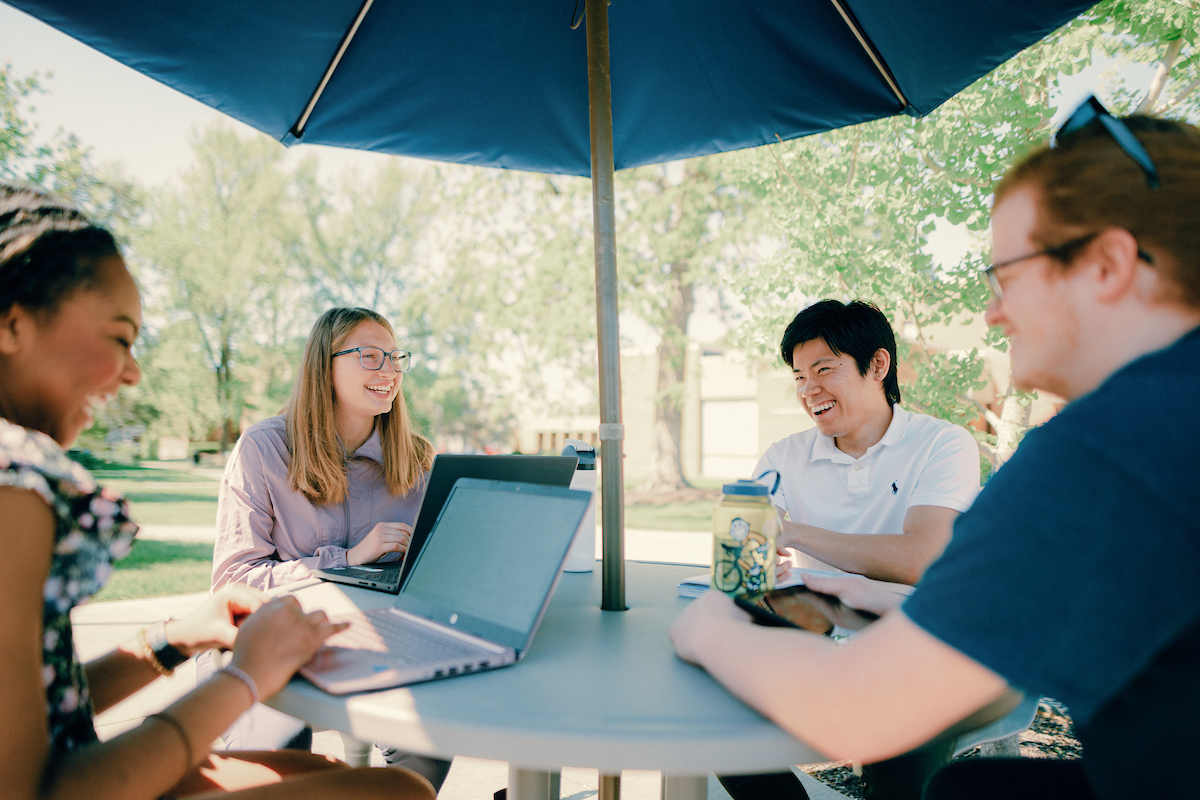 Four Columbia College students sitting around a patio table with umbrella on main campus, working on laptops and smiling during a discussion.