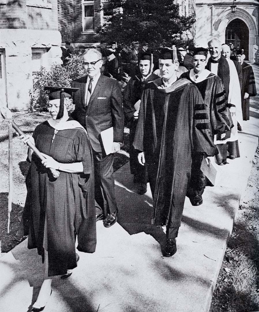 Historical image of male and female graduates and presenters walking toward graduation ceremony.