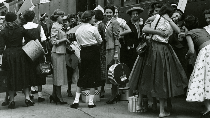 Historical image of a group of women, surrounded by luggage and hat boxes, saying goodbye while waiting in front of a train.