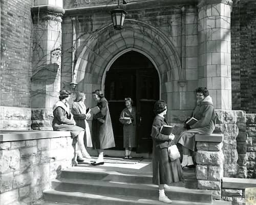 Historical image of a group of students wearing coats and sitting outside a main campus building on the stone ledge.