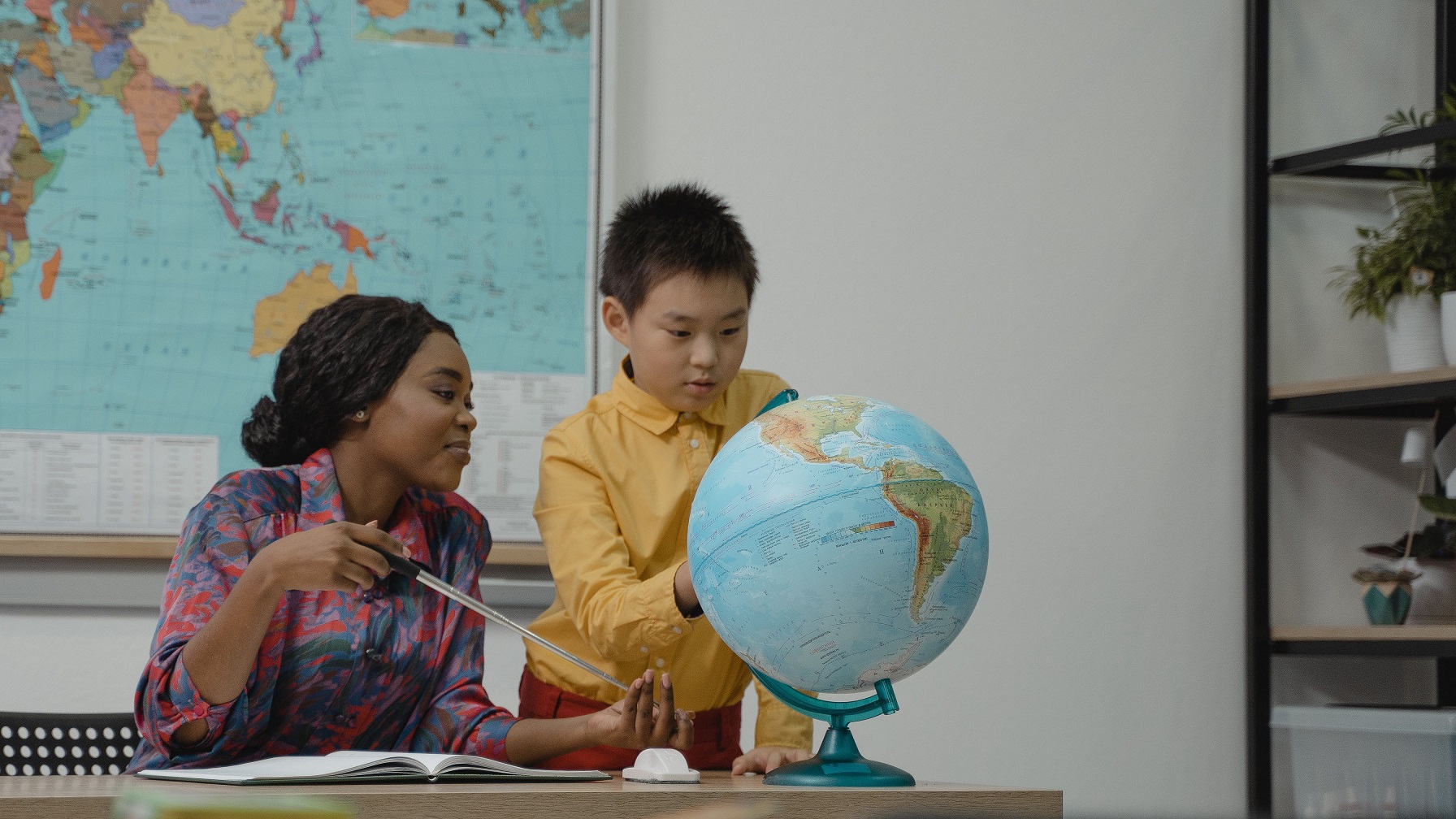  Teacher, sitting, and young student, standing, as they look over a world globe together.