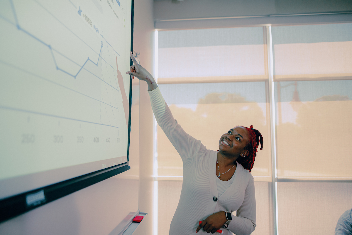 Woman in white dress standing in front of a window, pointing to a chart on the whiteboard.