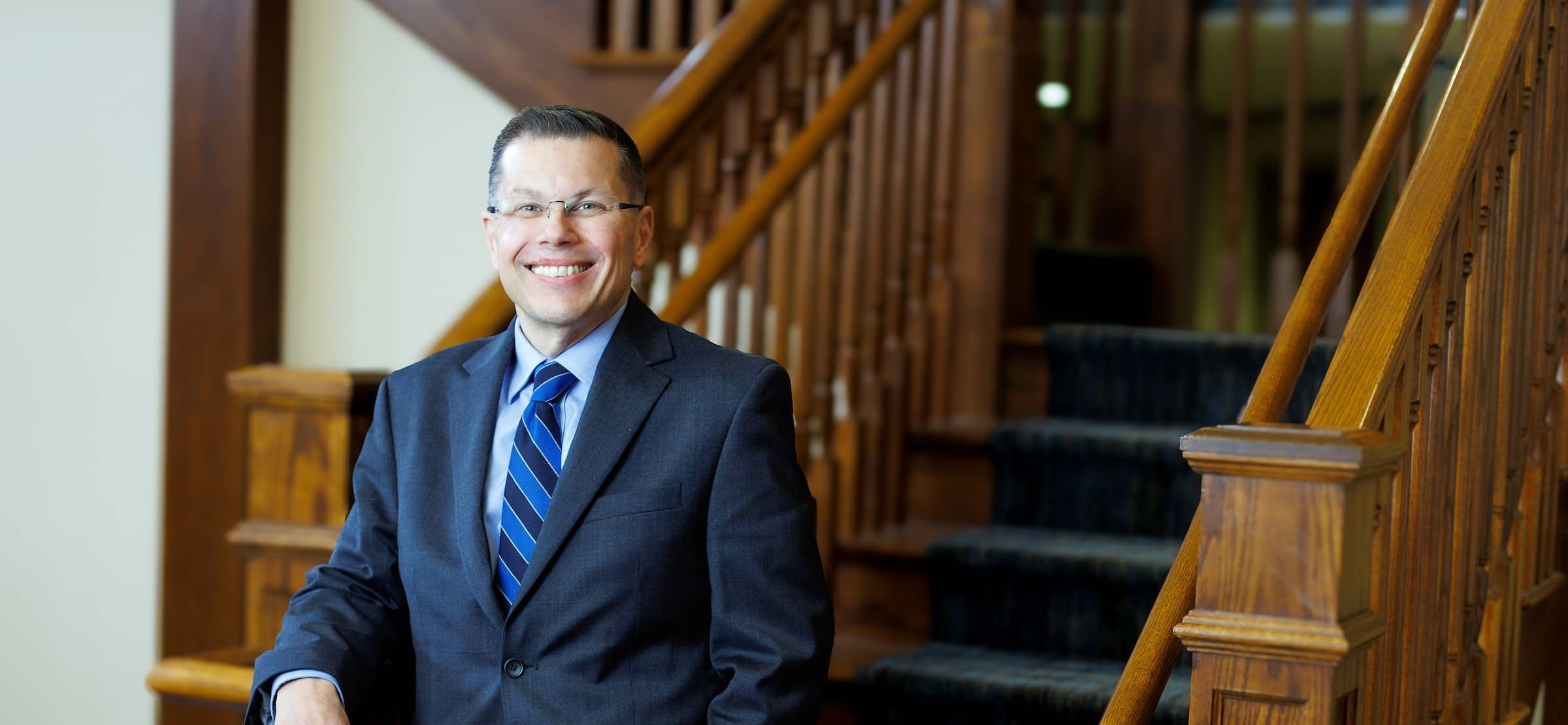 Dr. Toby Arquette, 19th President of Columbia College, wearing glasses, a dark suit and blue striped tie, standing in front of a staircase and smiling for the camera.