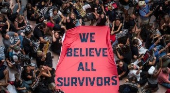 A large red sign being held by a group of people, saying "We Believe All Survivors".