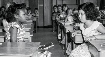 Two young girls sitting in a classroom having a conversation across the aisle.
