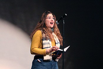 Student standing at a microphone reading from a book she is holding.