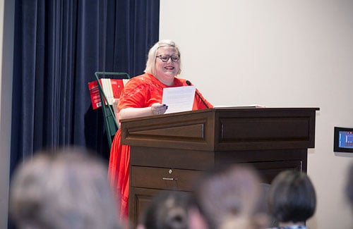 Woman in a red dress standing behind a large podium, reading from a piece of paper.