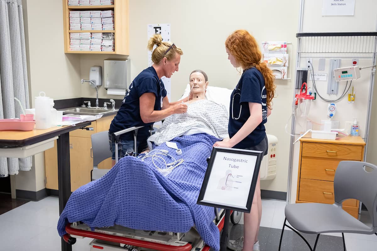 Instructor standing on left and nursing student standing on right of lab manikin.