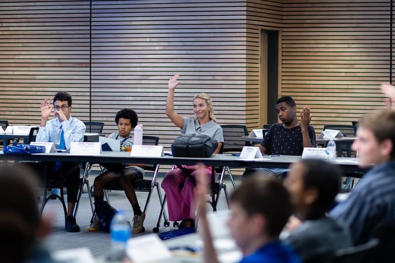 Four 2025 Voices of Leadership campers sitting at a table, name cards in front of them, three with their hands raised in response.