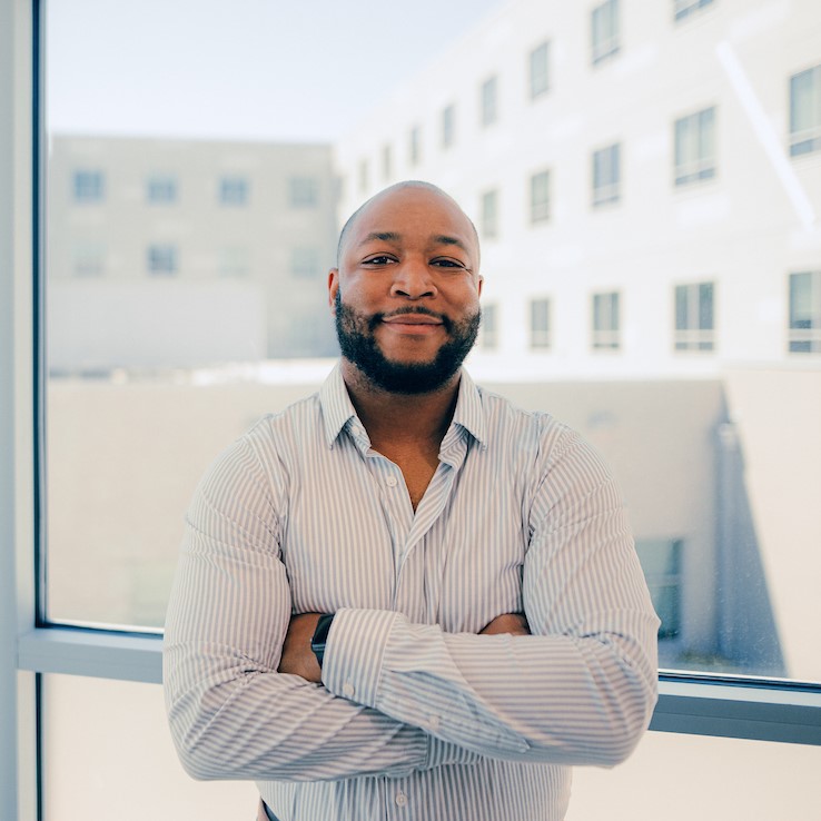 Graduate: Man standing inside in front of a window, smiling at the camera with arms crossed.