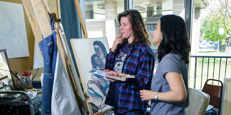 Student standing in front of her artwork on an easel, holding a paint palette, with her instructor standing next to her.
