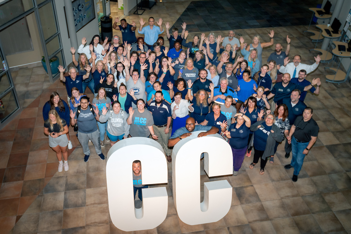Group of students, staff and faculty standing behind the large "CC" sign, waving at the camera.