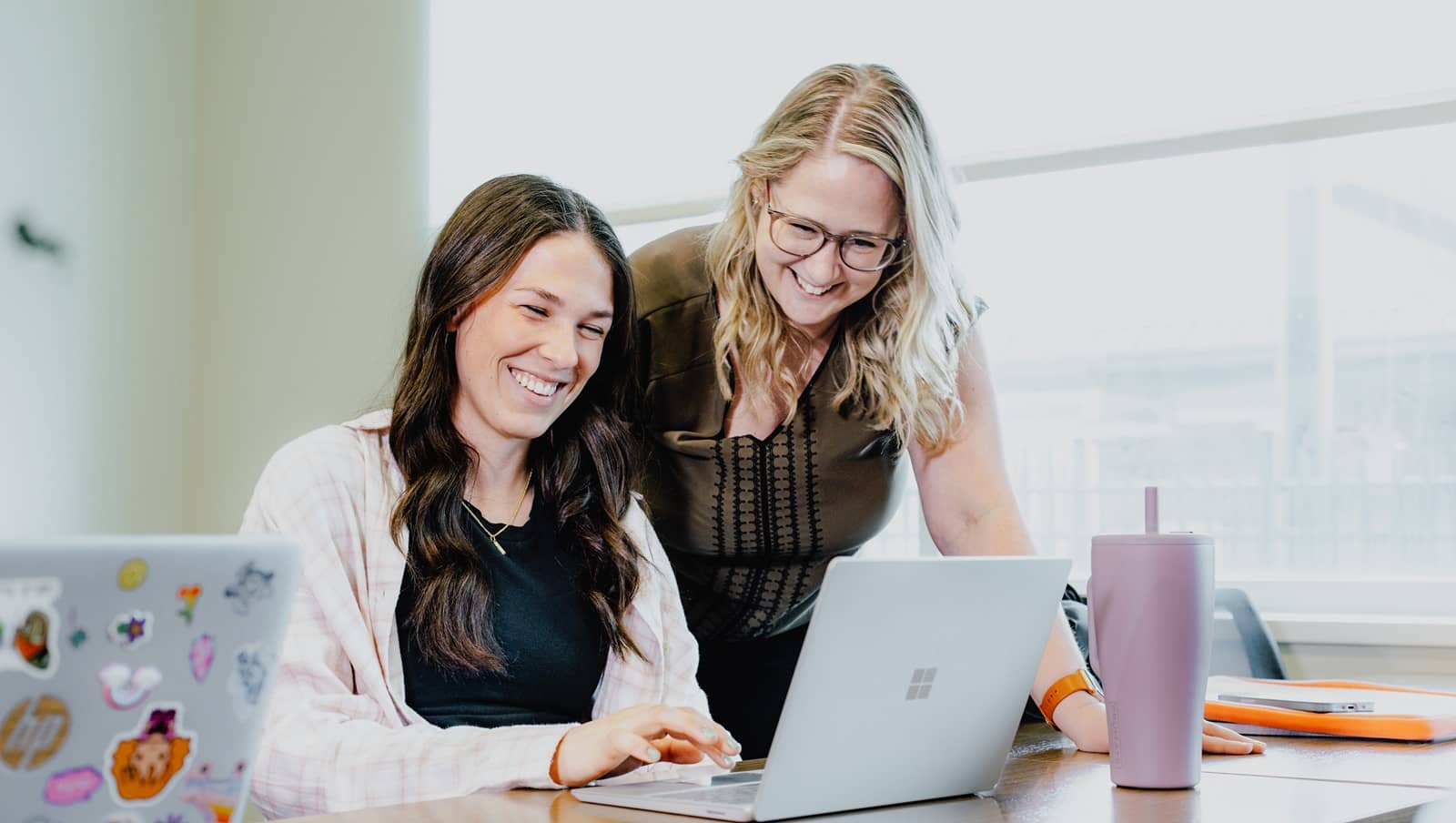 Director of CSMAC, standing, assists a student who is sitting at a table working on a laptop.