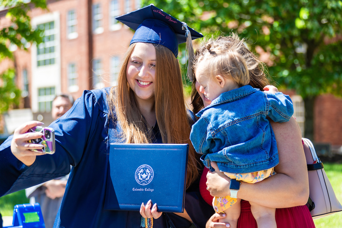 Picture of a female graduate in navy cap and gown, taking a selfie on her phone with a young child being held by another woman.