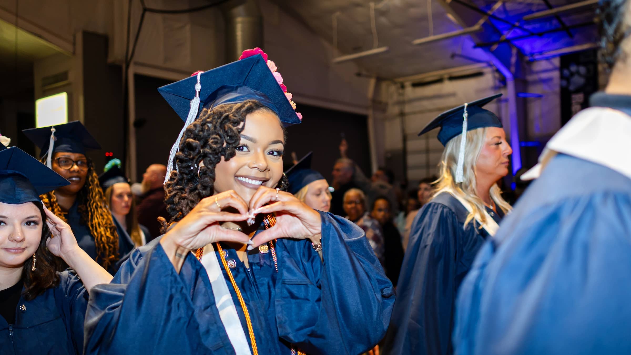 Smiling female graduate making a heart shape with her hands, surrounded by other CC graduates.