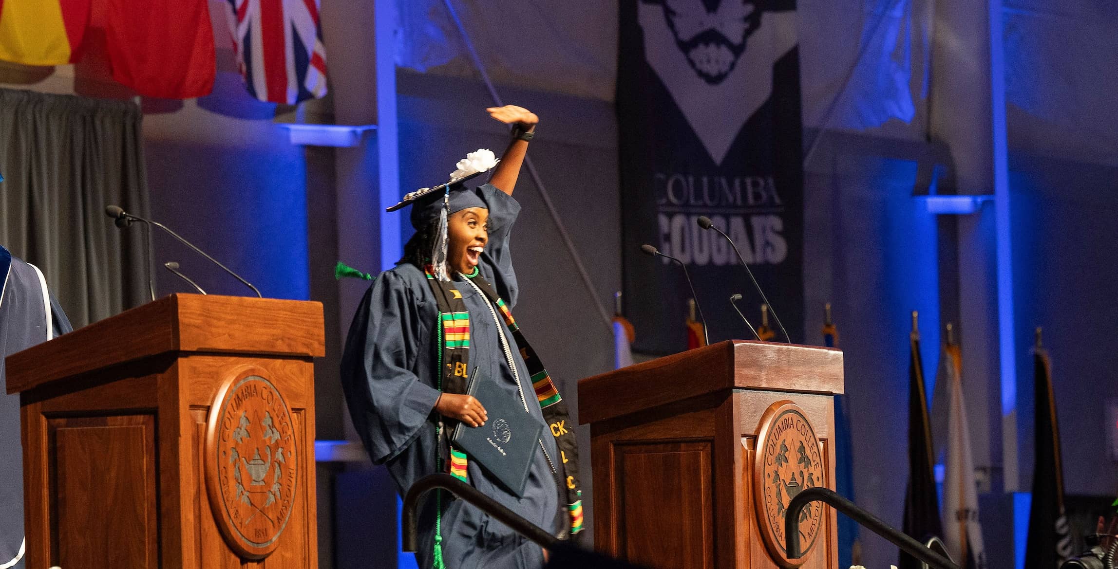 Spring 2025 CC graduate celebrating her achievement at the commencement ceremony, with joyous smile on her face and left hand in the air.