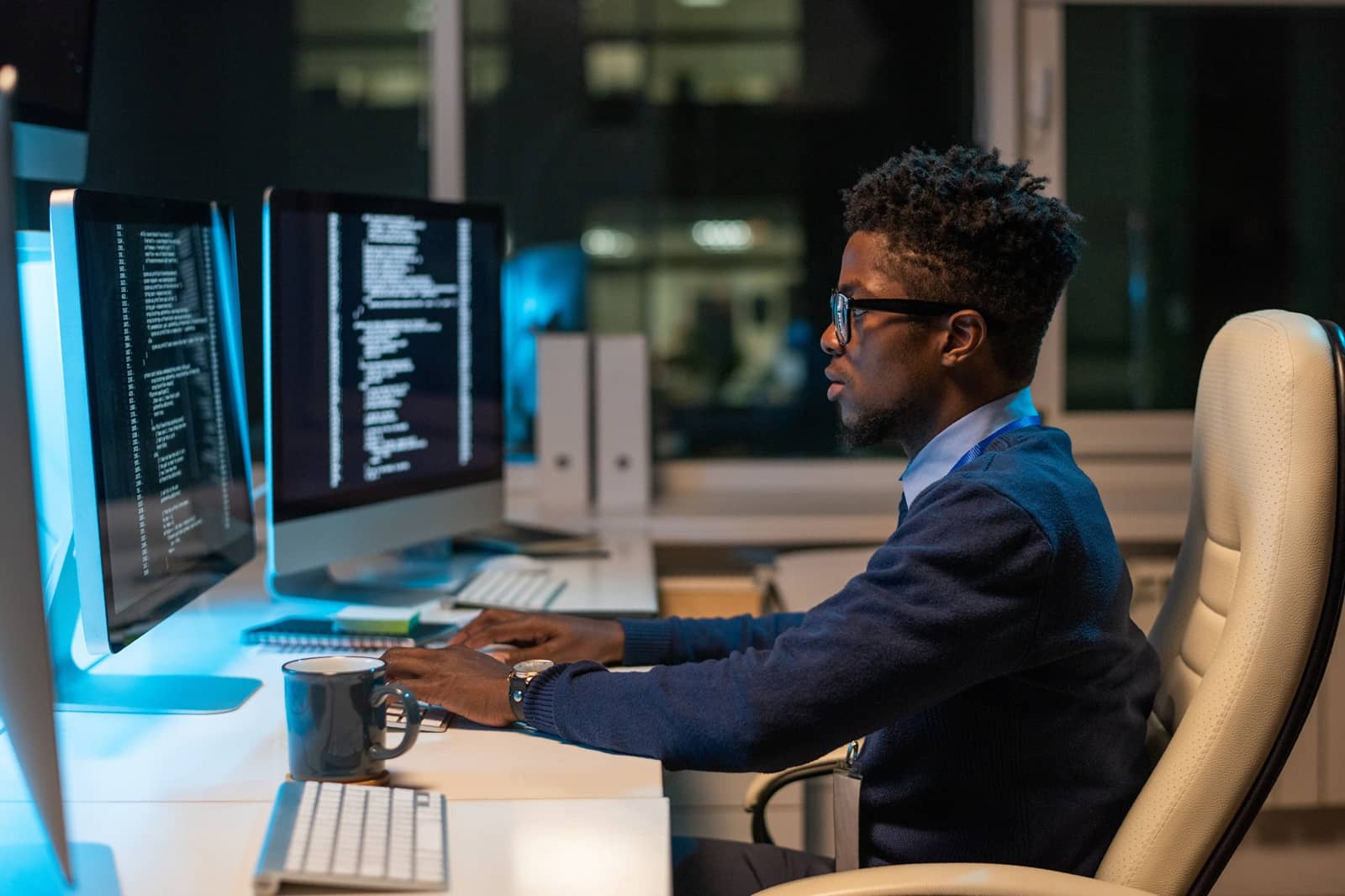 Young professional of African ethnicity using his Master of Science in Cybersecurity degree by reviewing coded data while sitting in an armchair in front of computer in office.