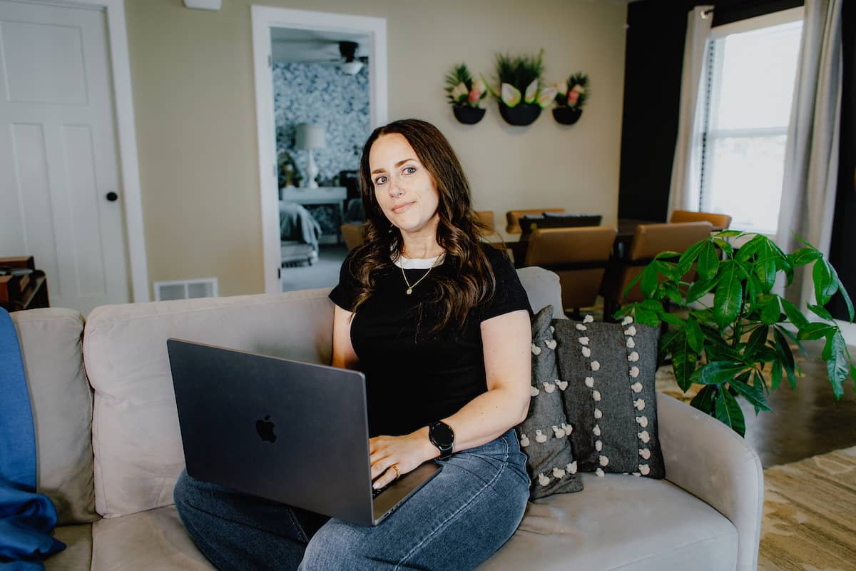 Female student, wearing a black t-shirt and jeans, sitting on her coach with a laptop in her lap.