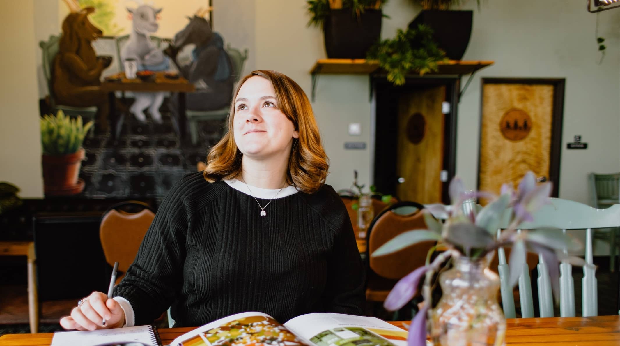 Female adult student with auburn hair, wearing a dark sweater, and sitting at a table studying in a coffee shop.
