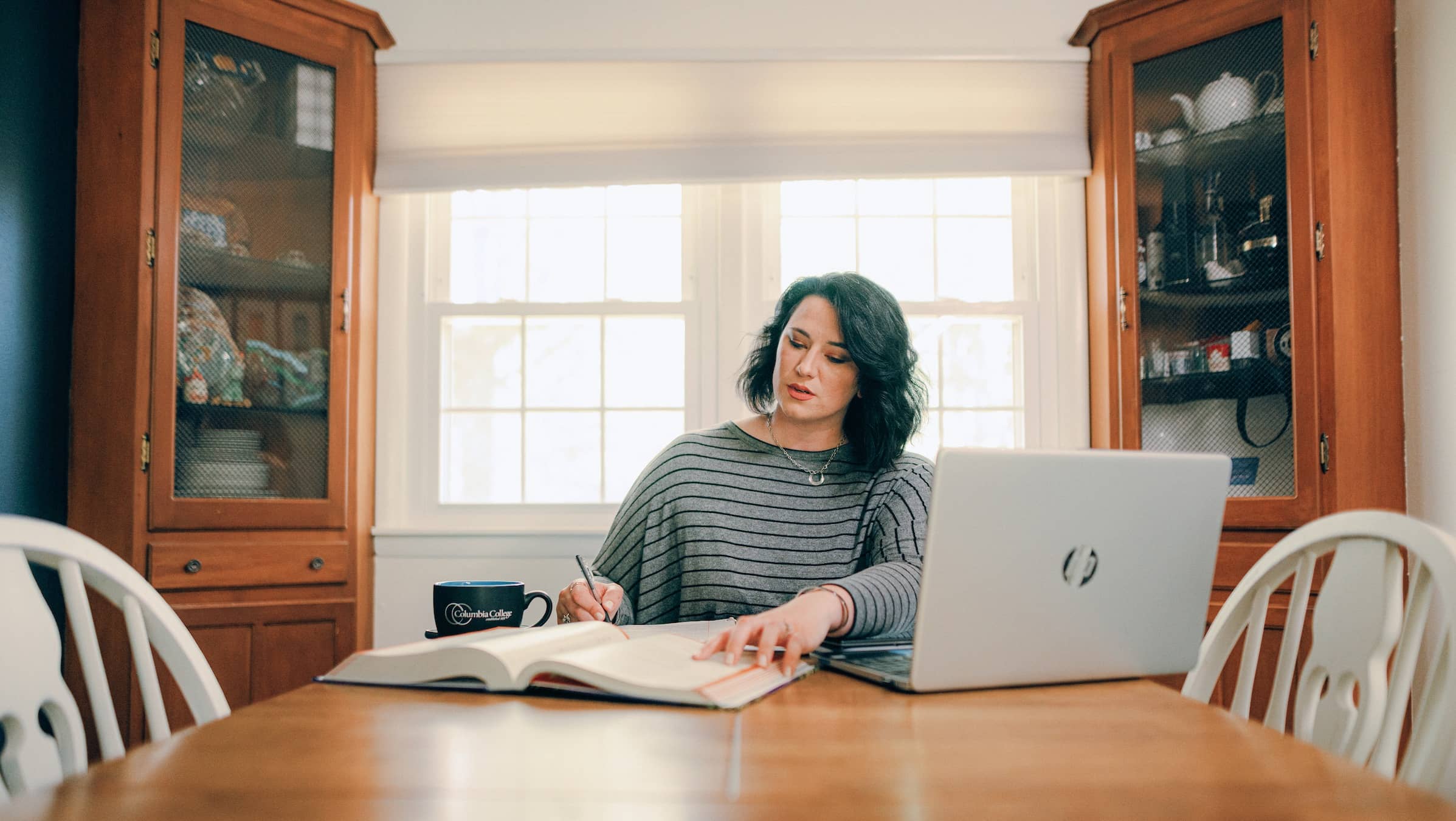 Female adult student, wearing a gray striped shirt, sitting at her dining table and taking notes from a book, with a laptop in front of her.