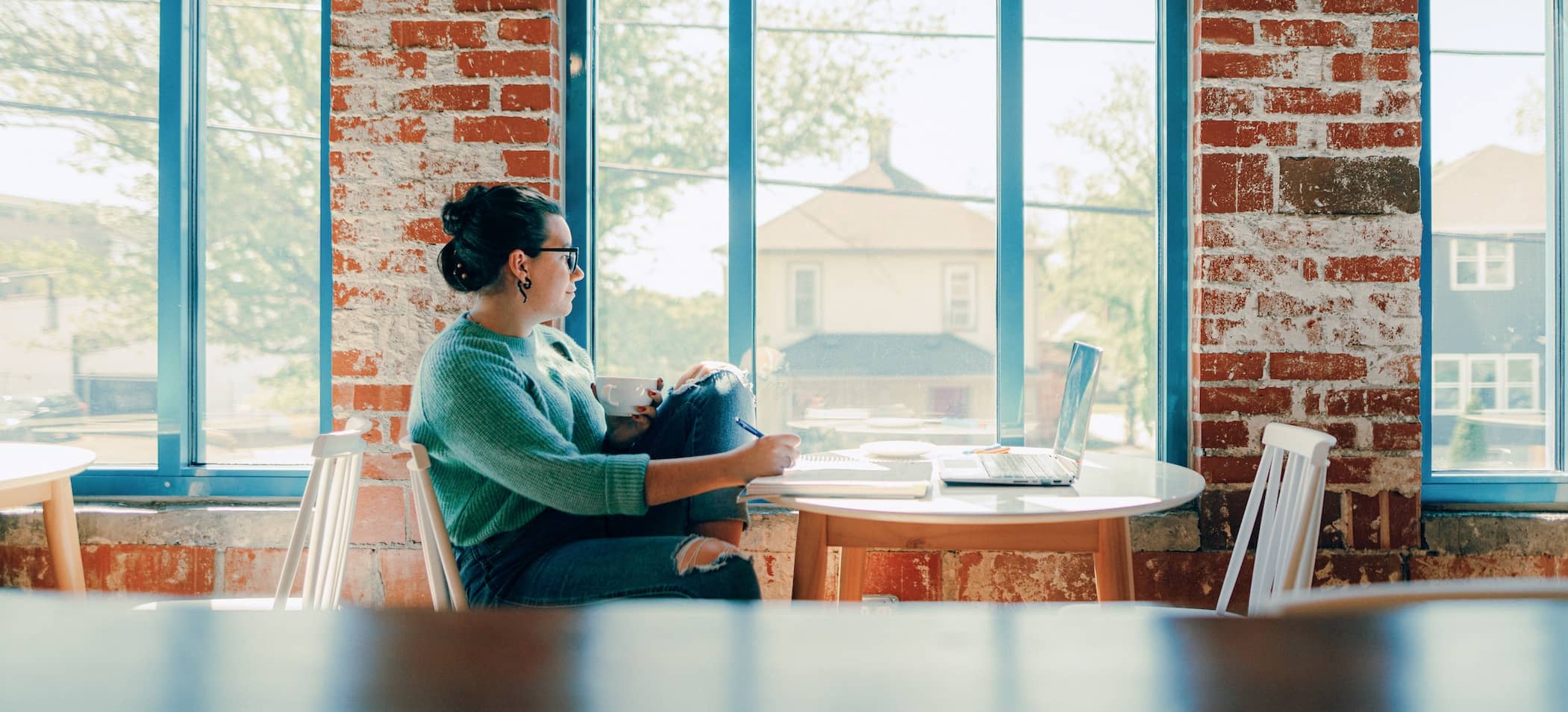 Female adult student, wearing glasses, a green sweater with her hair pulled up, sitting at a table in a coffee shop in front of a notepad and laptop.