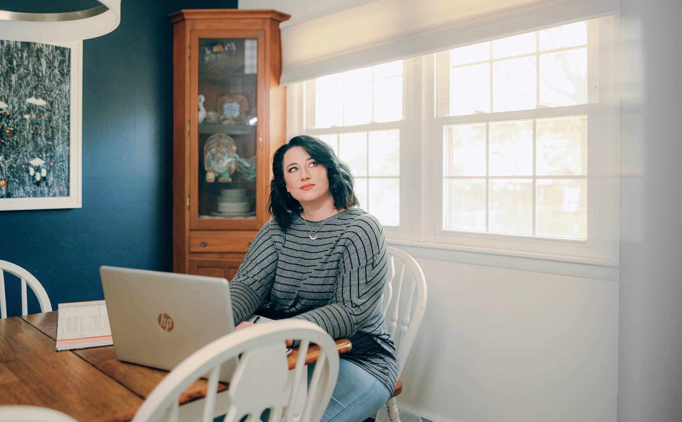 Adult student, sitting at a dining table with a laptop in front of her and a window full of sunshine behind her.