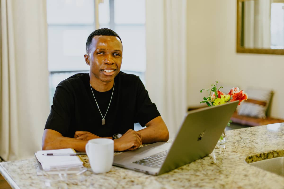 Male student, wearing a black t-short and sitting at kitchen counter with a laptop, cup of coffee, and notepad in front of him.