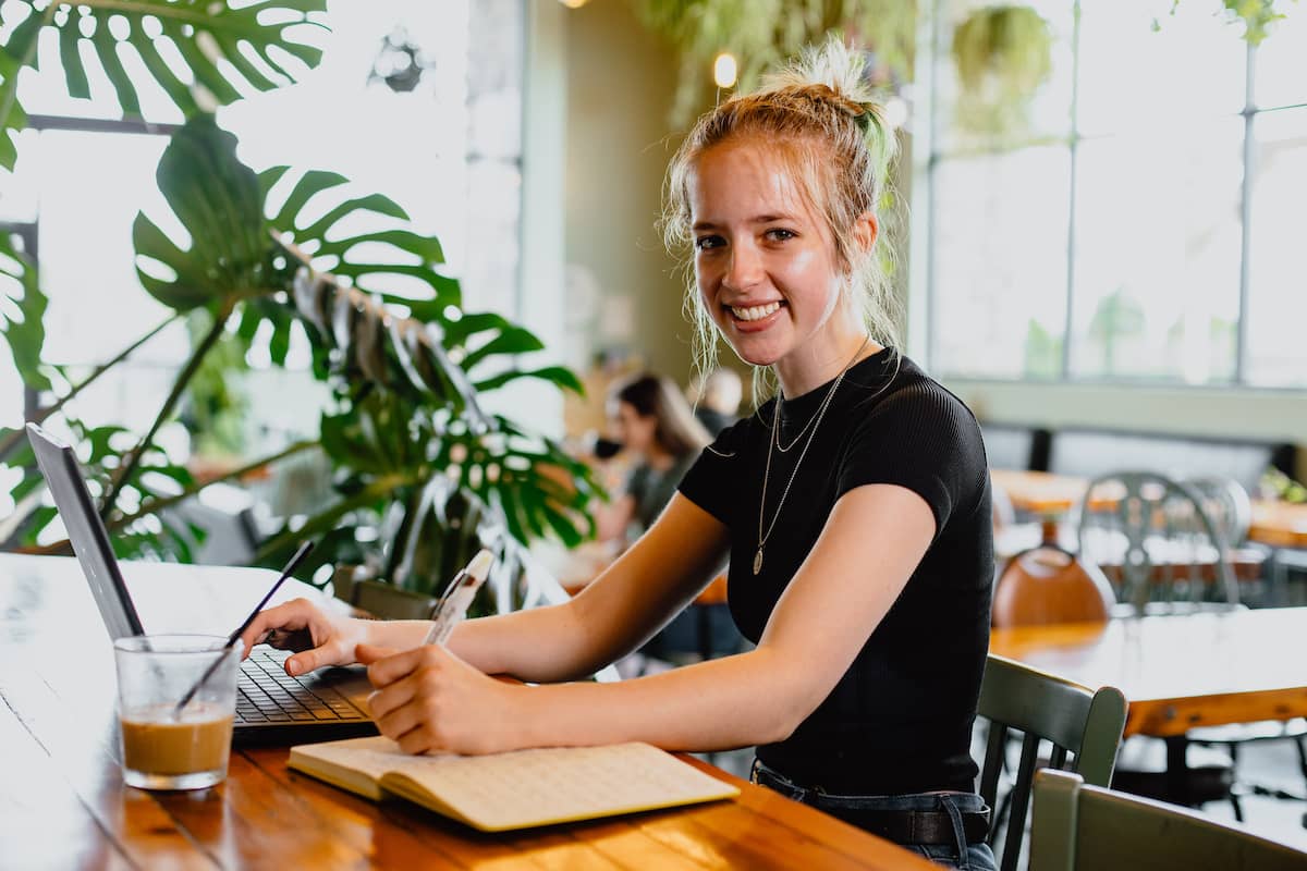 Female student, wearing black t-shirt, sitting in a coffee shop studying, with a laptop, book, and a coffee in front of her.