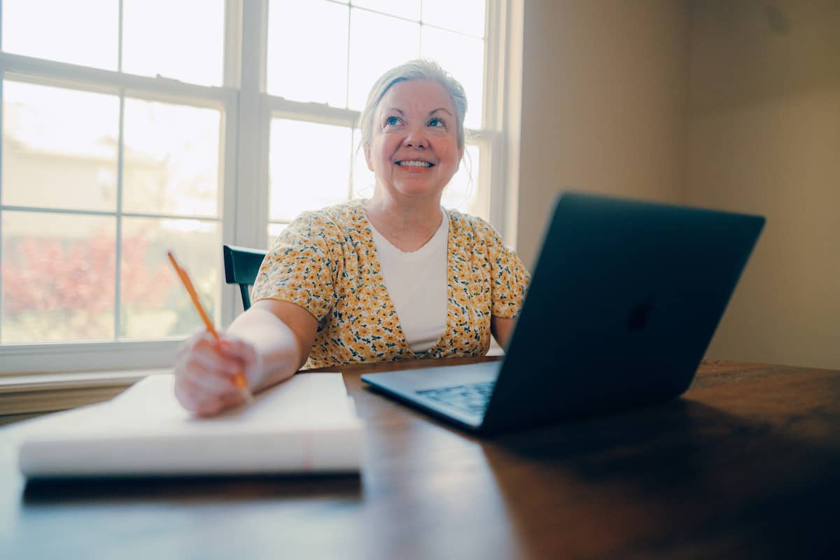 Adult student sitting at dining table in front of window, with a laptop and writing pad in front of her.