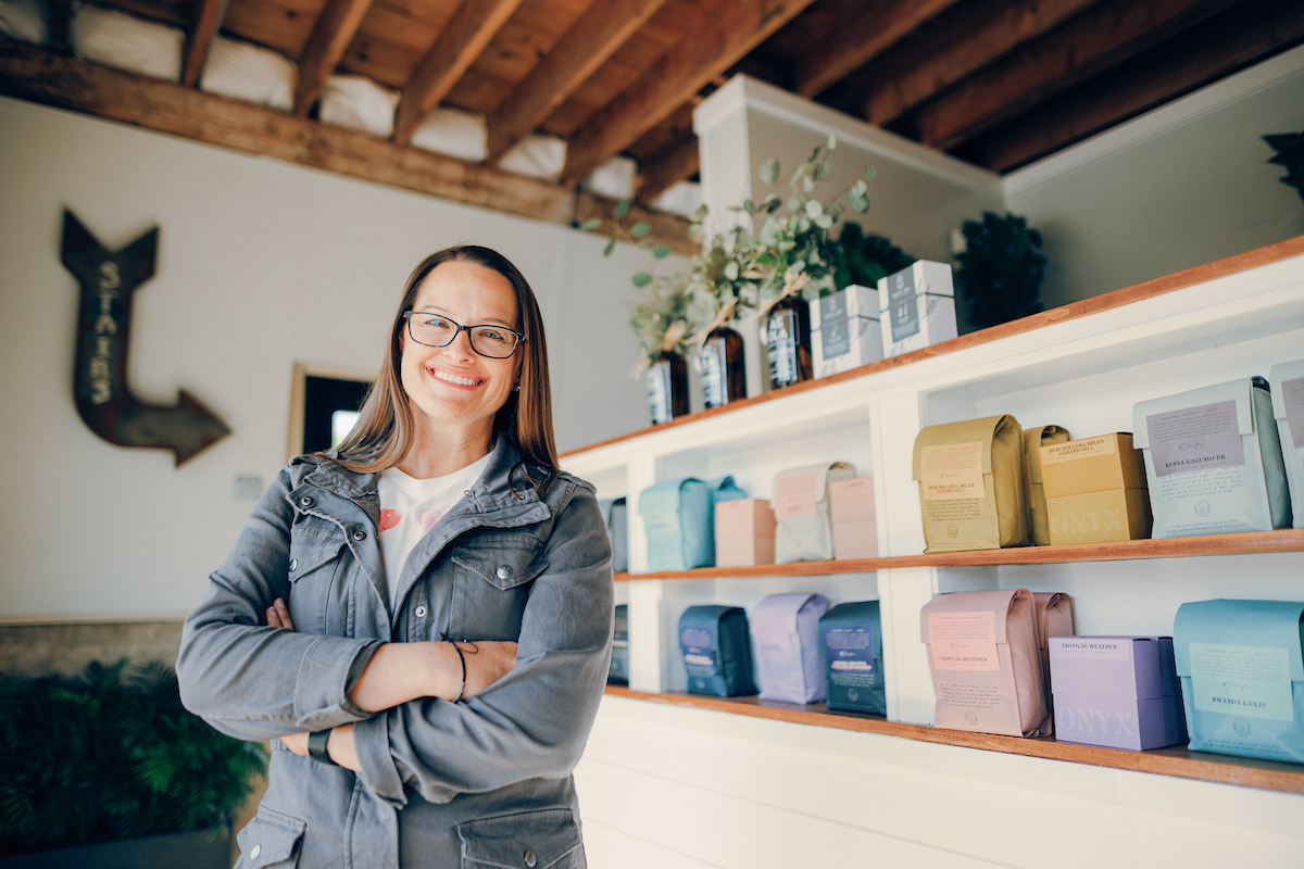 Female business owner, wearing glasses, smiling at the camera while standing in front of shelves displaying her products.