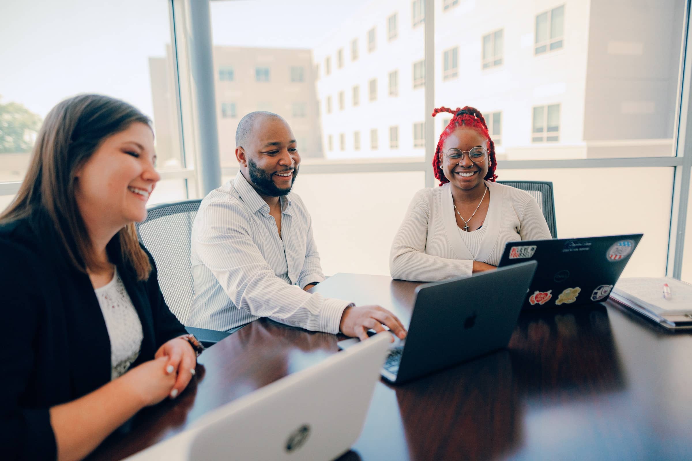 Two female and one male public administration business professionals in conversation while sitting at a table, with a laptop in front of each.