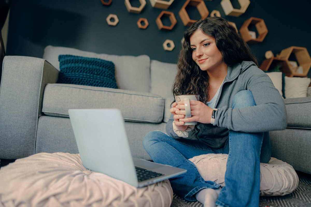 Syllabi: Woman sitting on a couch holding a coffee mug while looking at a laptop.