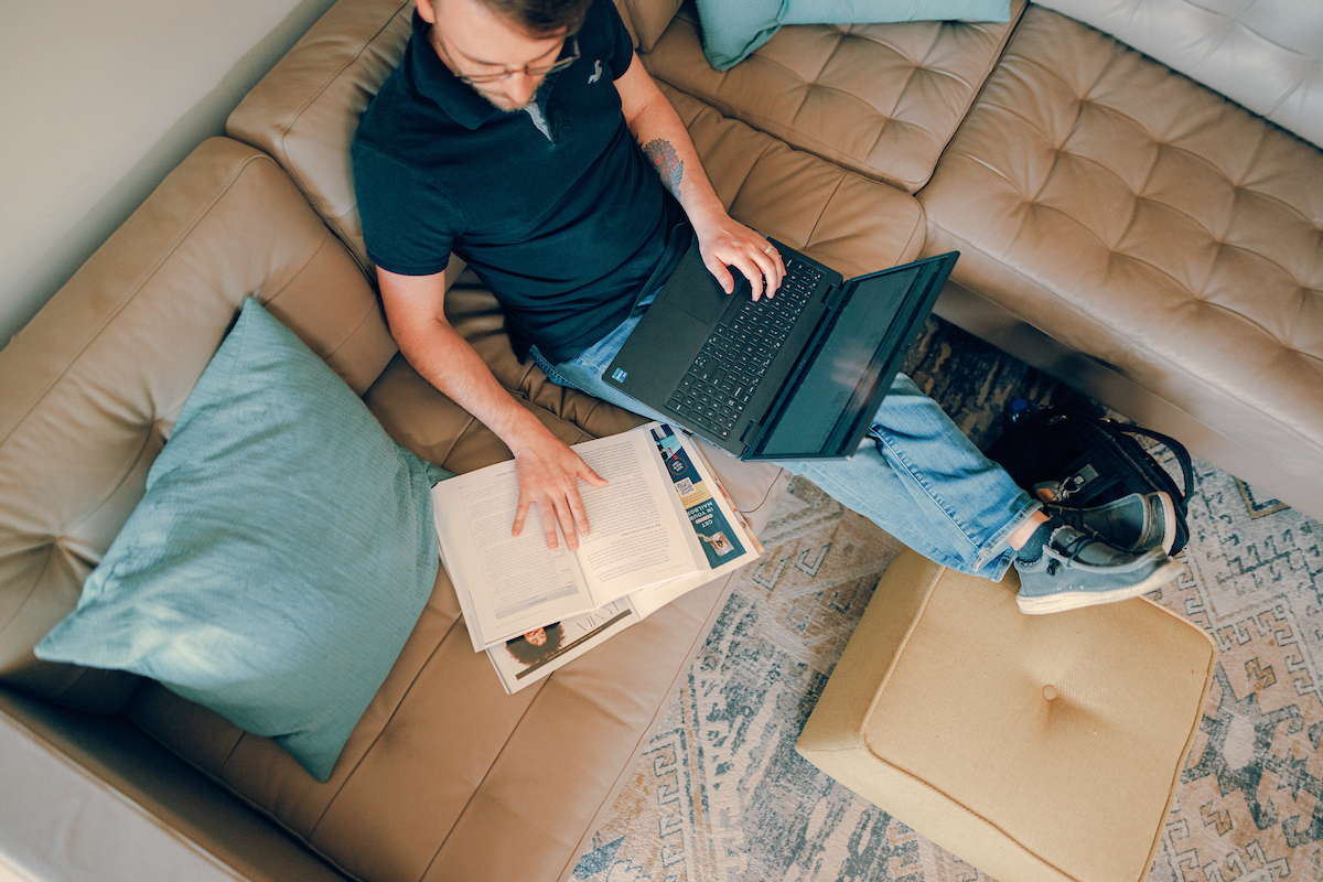 Overhead view of man sitting on a couch, feet propped on an ottoman, reviewing documentation and working on a laptop.
