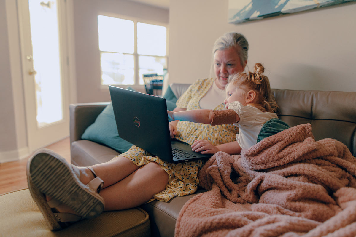 Online student sitting on a couch with her granddaughter looking at her laptop.