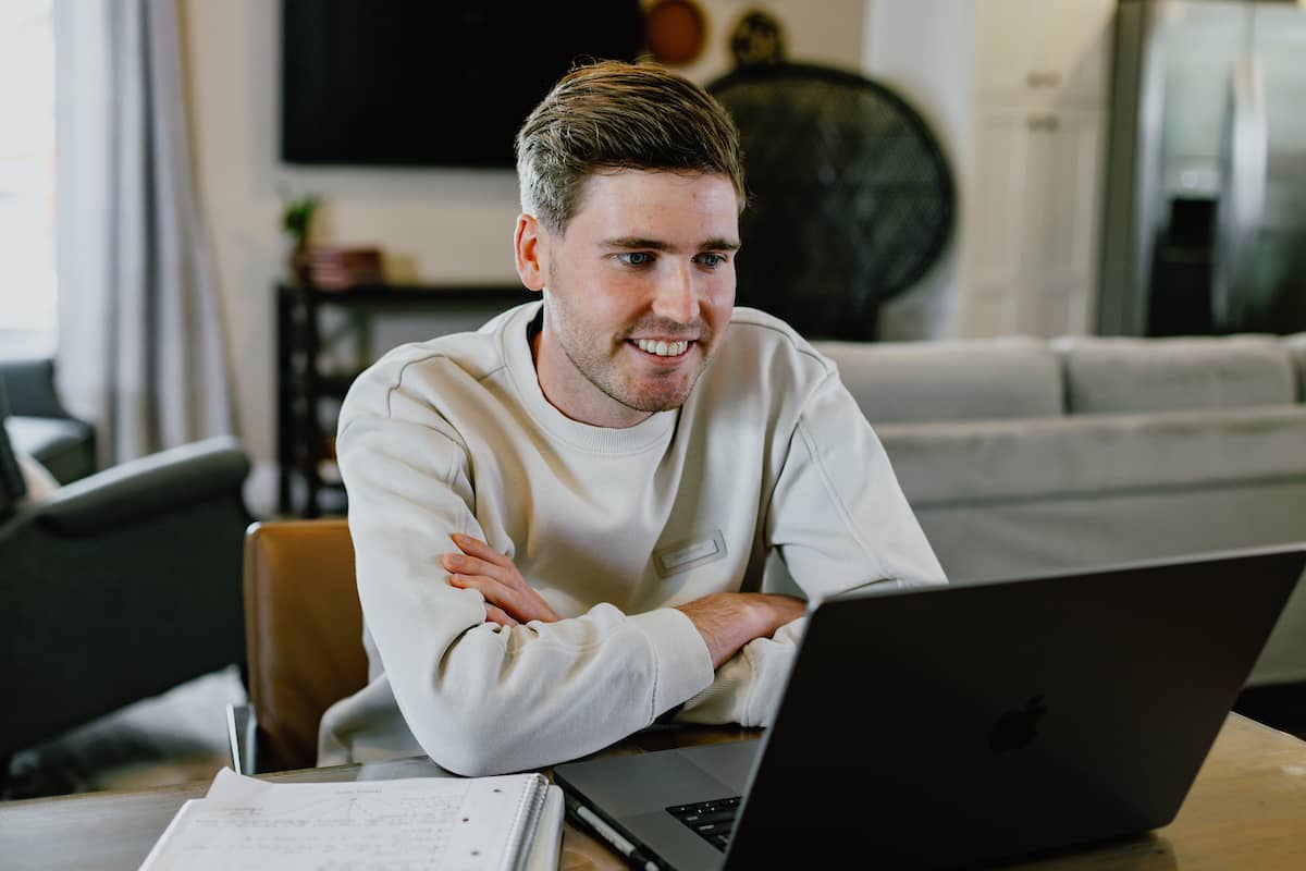Man wearing a long sleeved white shirt, sitting at a table in his home, with a notebook and laptop in front of him.