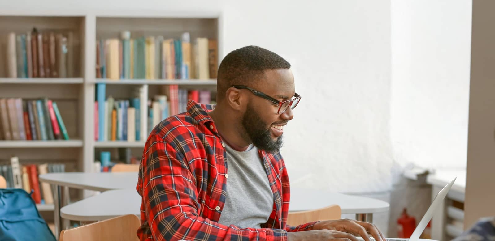 Male online master's program student, typing on his laptop and wearing a red flannel shirt and glasses.