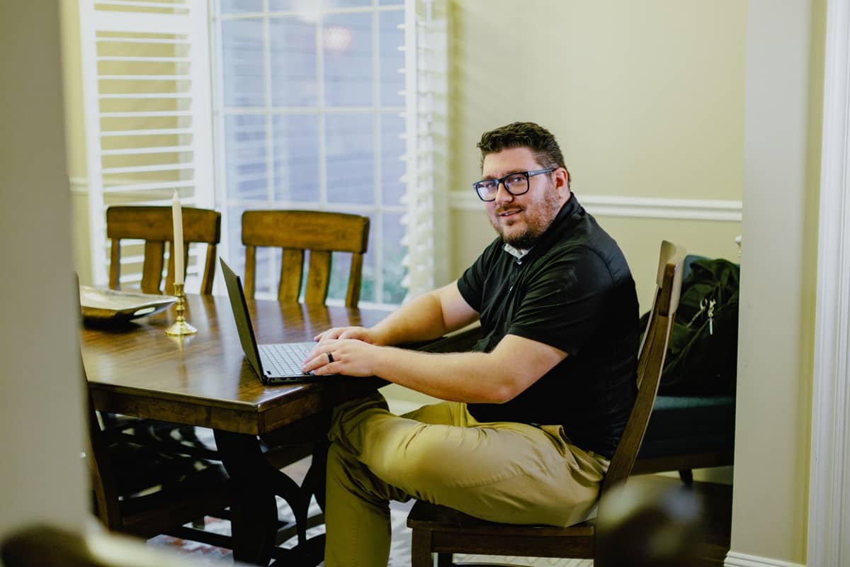 Master of Public Administration student wearing a black polo and khaki pants, sitting at his kitchen table working on a laptop.