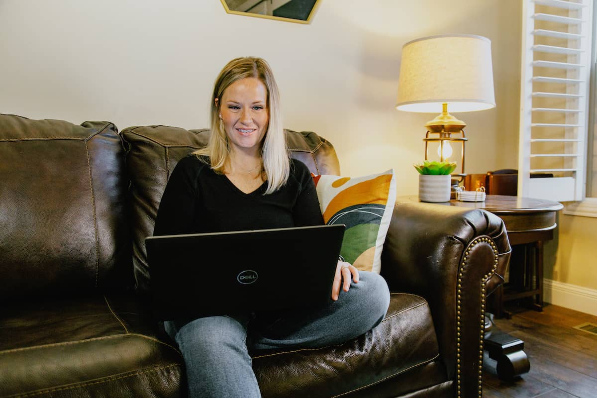 Female adult student with blonde hair, wearing a black shirt and sitting on her leather couch, working on her laptop.