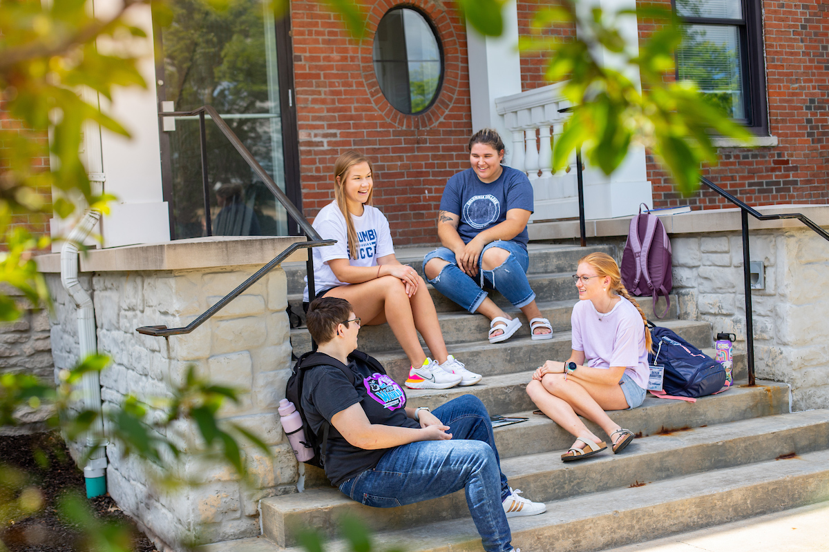 Group of four students sitting on the entry steps of a brick building on a sunny day.