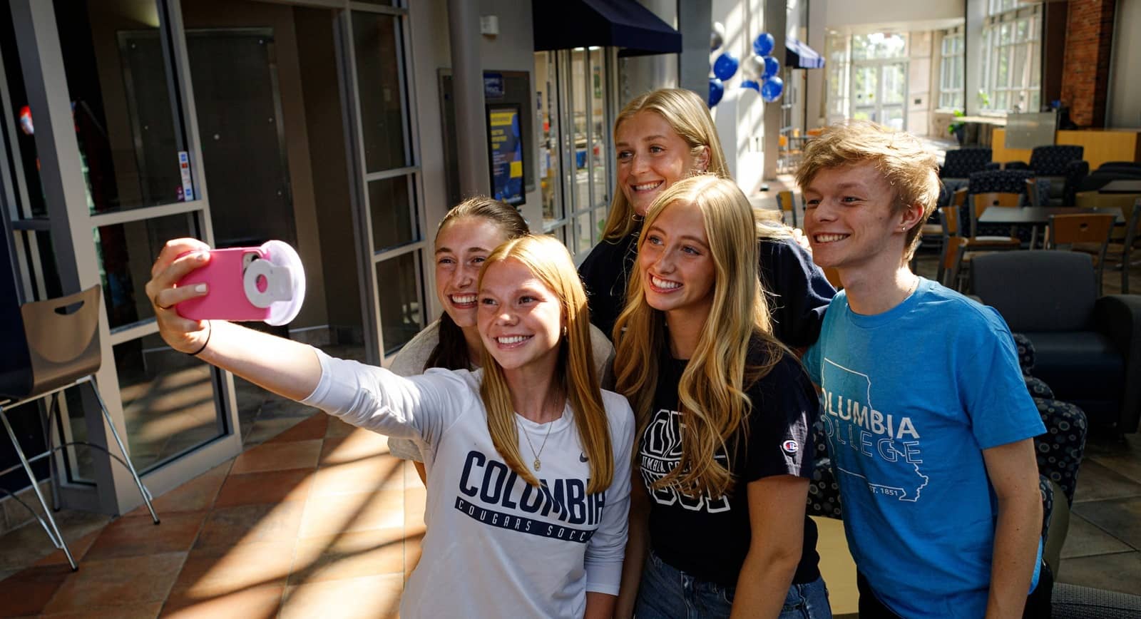 Five students standing inside on main campus, posing together for a selfie, all sporting Columbia College shirts.
