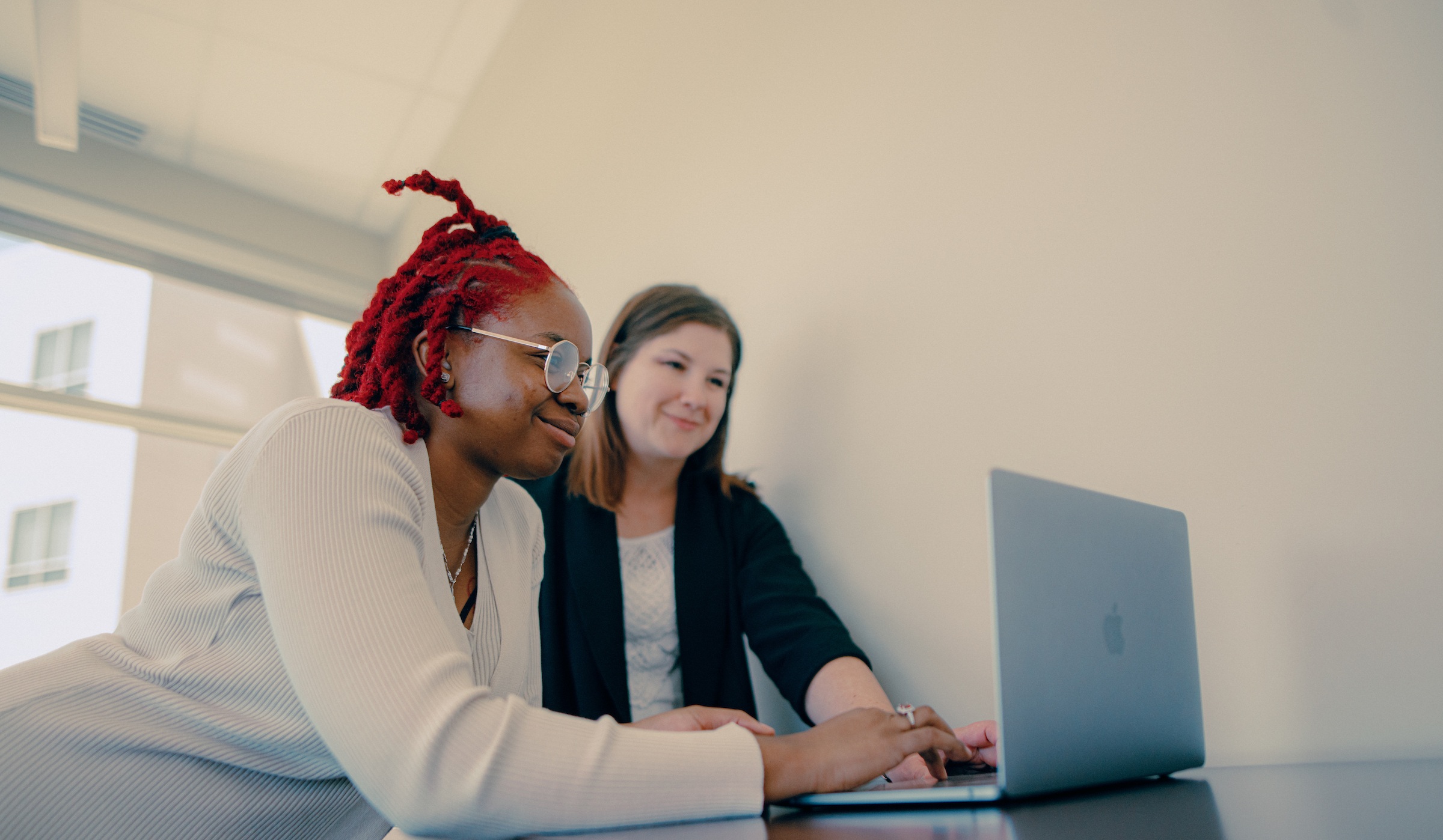 Two female business leaders, one wearing glasses and a white sweater and the other wearing a black sweater, reviewing content on a laptop.