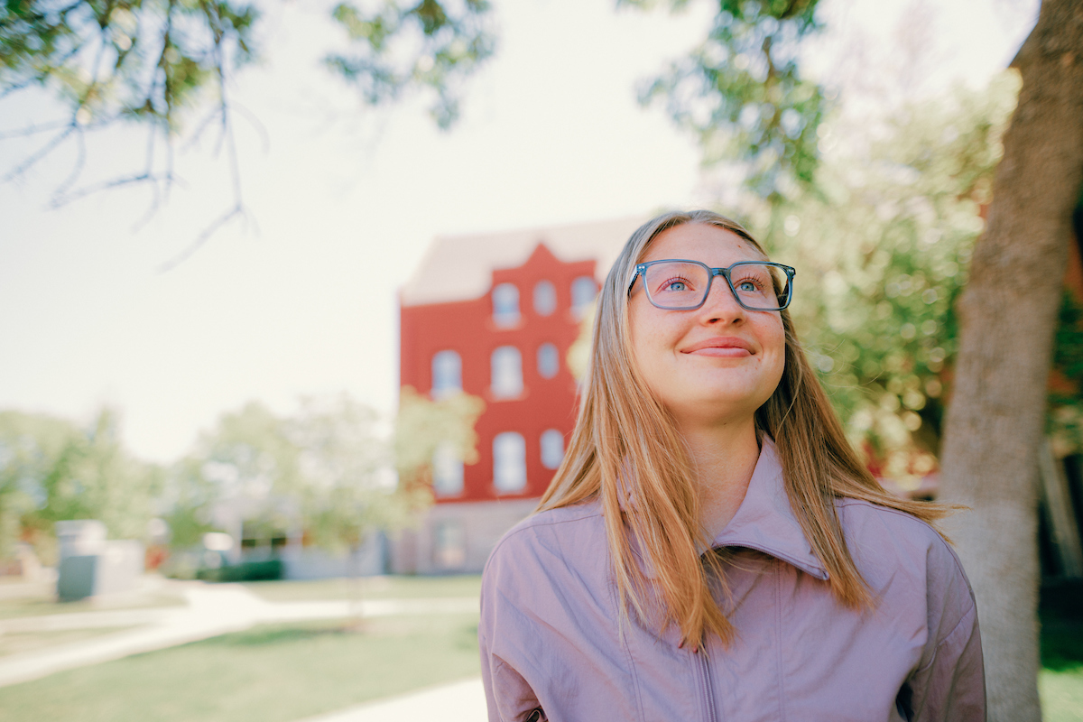 Student, wearing glasses, standing outside on main campus and staring up at the sky.