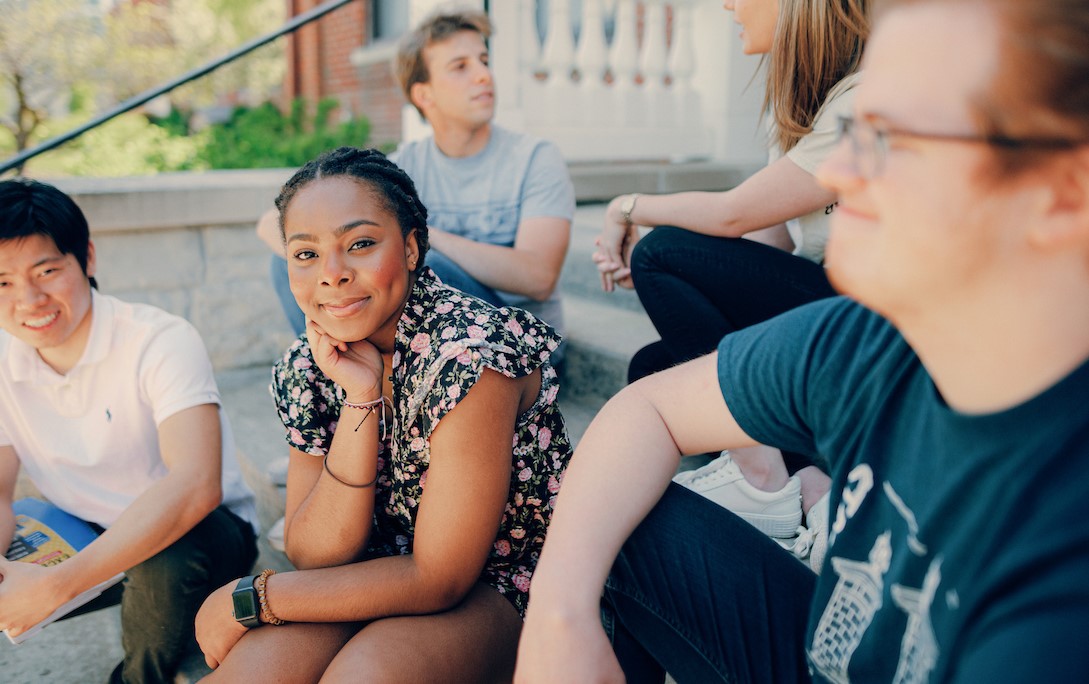 Female student sitting on cement steps, surrounded by other students, with her chin resting on her hand while she smiles directly at the camera.