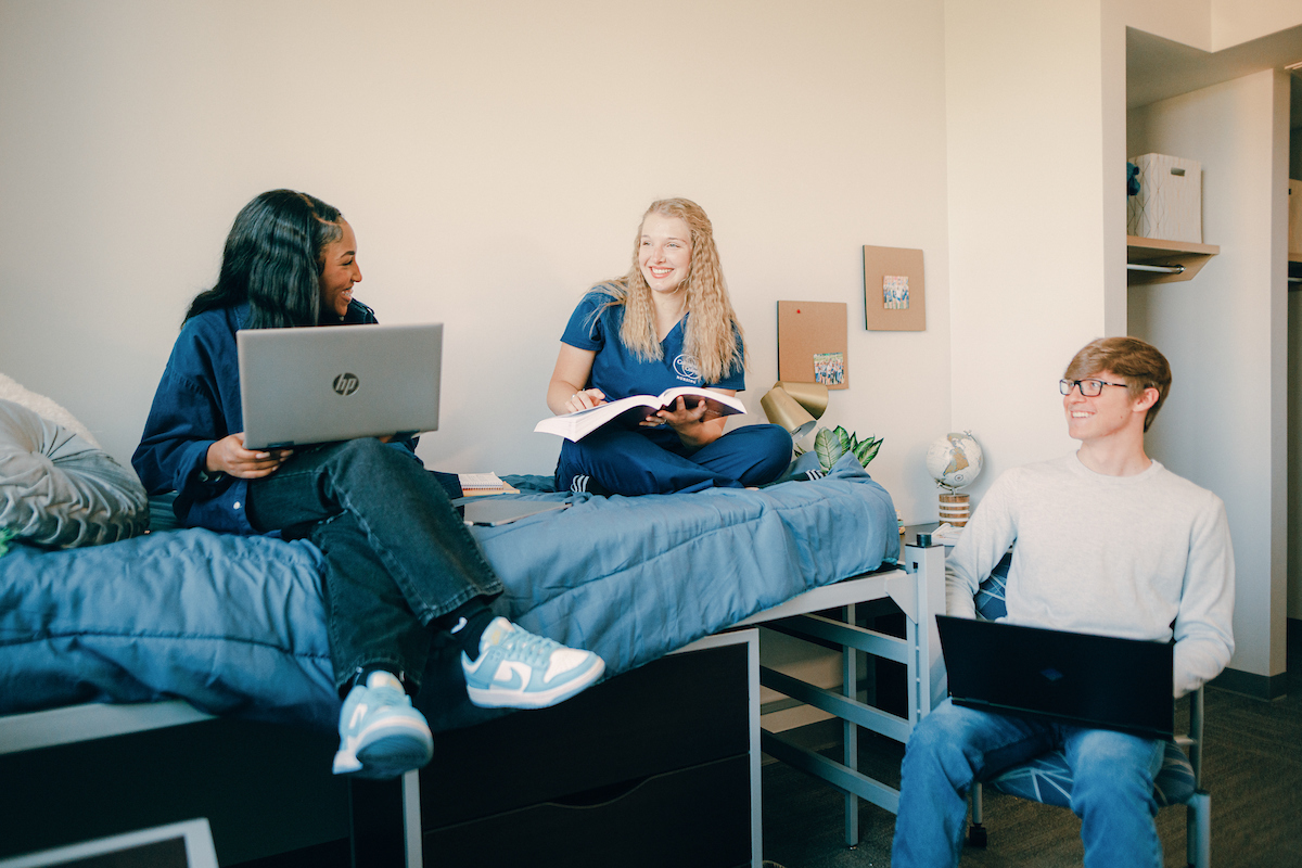 Three students in a residential hall room, two seated on bed with a laptop and textbook and one seated in chair with a laptop.